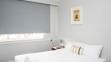 A hotel room with a bed, a nightstand, a lamp, a framed picture on the wall, and a window with a blind.