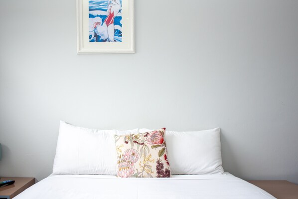 Una cama bien hecha con ropa blanca y una almohada estampada con flores. Un cuadro enmarco cuelga de la pared sobre la cama.
