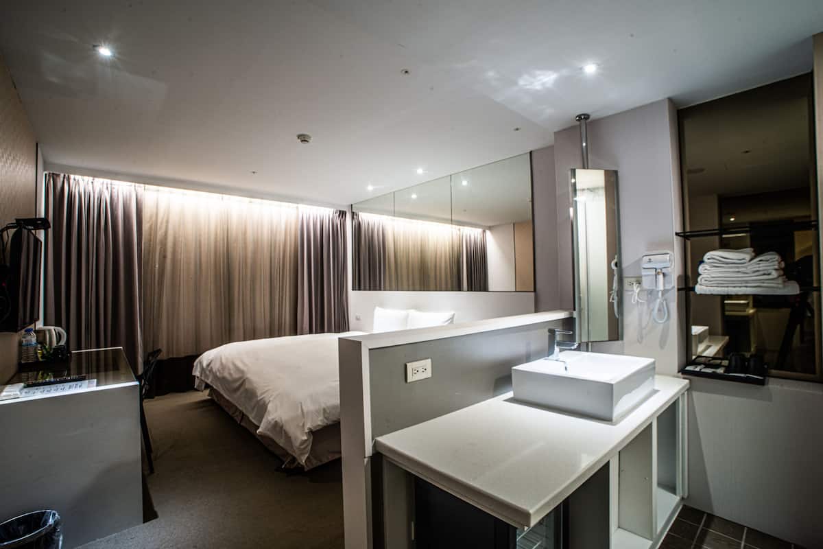 A modern hotel room with a single bed, a large mirror, a built-in sink, and a small desk with a trash bin.