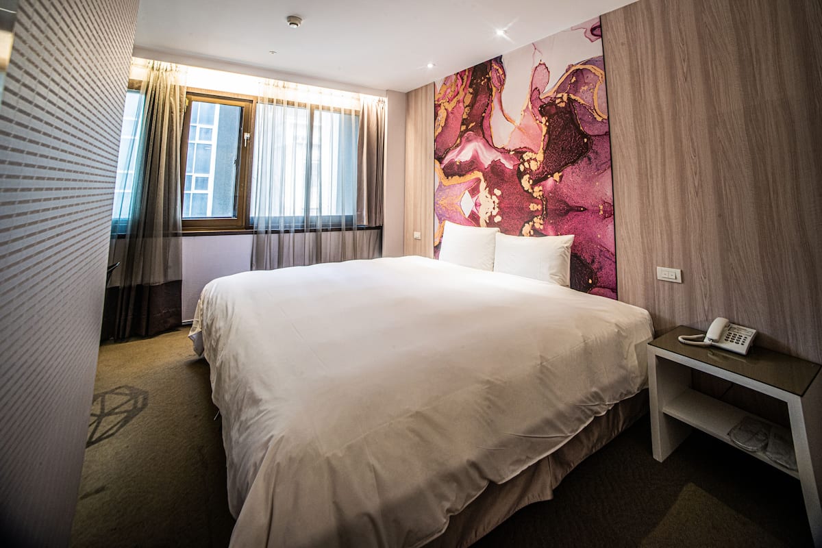 A hotel room with a large bed, a bedside telephone, and a window with curtains.