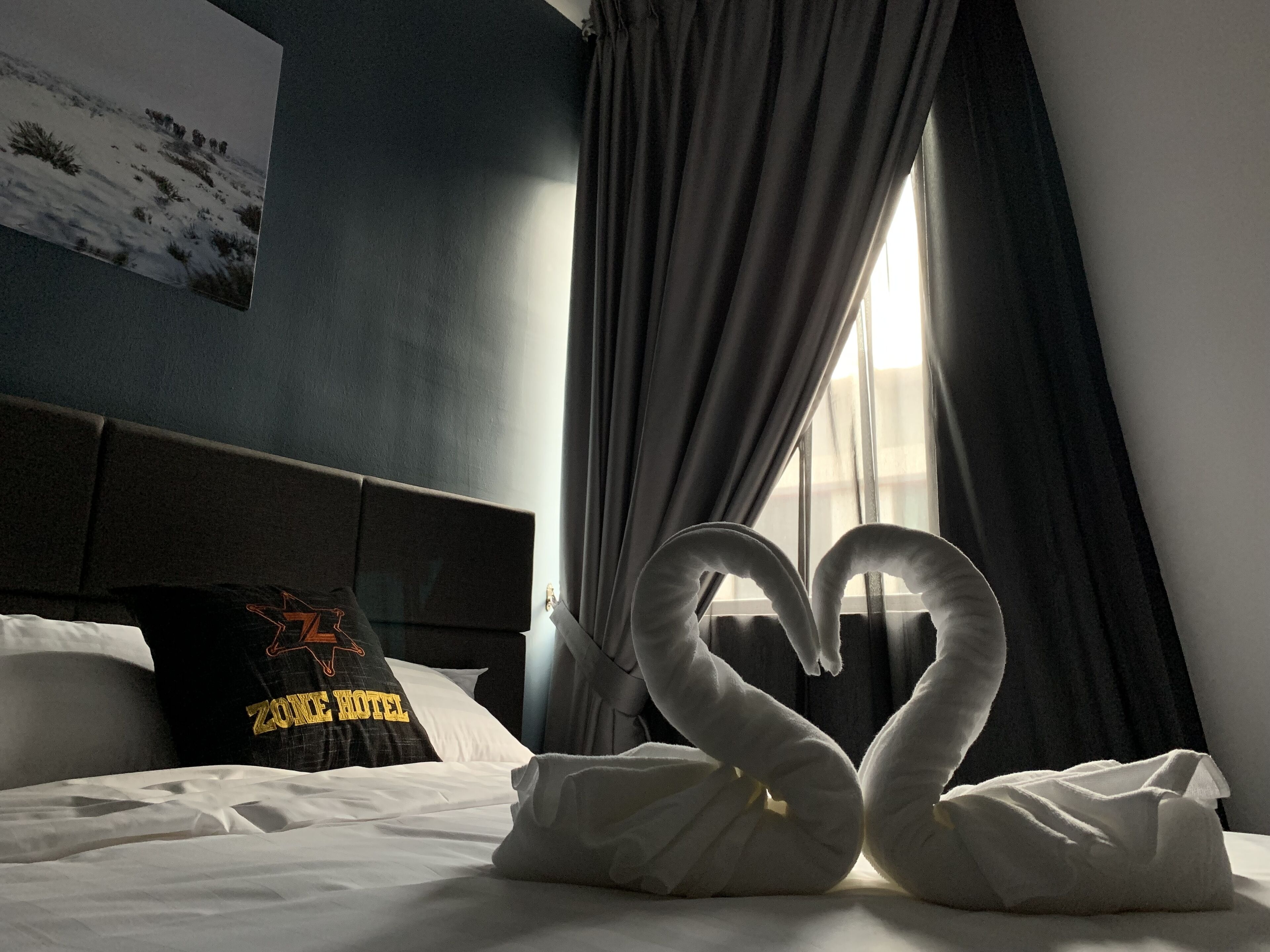 A hotel room with a bed, a pillow with a logo, a window with curtains, and a swan-shaped towel arrangement.