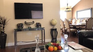 A living room with a TV mounted on the wall, a glass coffee table with a fruit bowl, a vase, and a remote control. - POOL/JACUZZI/HUGE FURNISHED BACKYARD/POOL TABLE/MINI GOLF/PLAY PLACE/PING PONG T (Las Vegas)