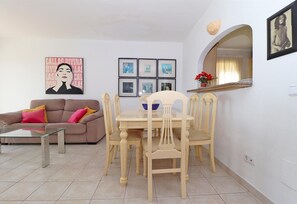 Un salon comprenant un canapé, une table basse en verre, une table à manger avec des chaises, des tableaux encadrés accrochés au mur et une fenêtre avec un miroir.