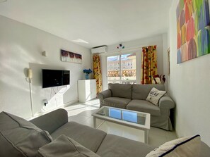 Un salon comprenant un canapé, une table basse, un téléviseur à écran plat, un meuble et une fenêtre avec des rideaux.