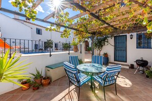 A patio with a round glass table and striped chairs, surrounded by potted plants and a pergola with vines.