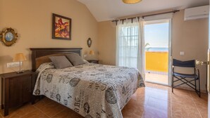 Un dormitorio con cama, mesita de noche, lámpara, cuadro, ventana con cortinas y vistas al exterior.