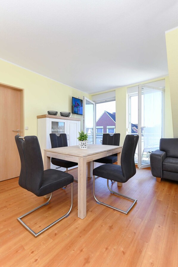 A dining area with a wooden table, four black chairs, a white cabinet, and a TV on the wall. - Vacation apartment Flut - Holiday apartments in Haus Seekieker in Bensersiel (Esens)