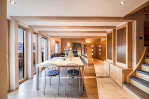Un vaste espace dédié à la salle à manger, avec une longue table et des chaises, des poutres en bois au plafond et de grandes fenêtres offrant une vue sur l’extérieur.
