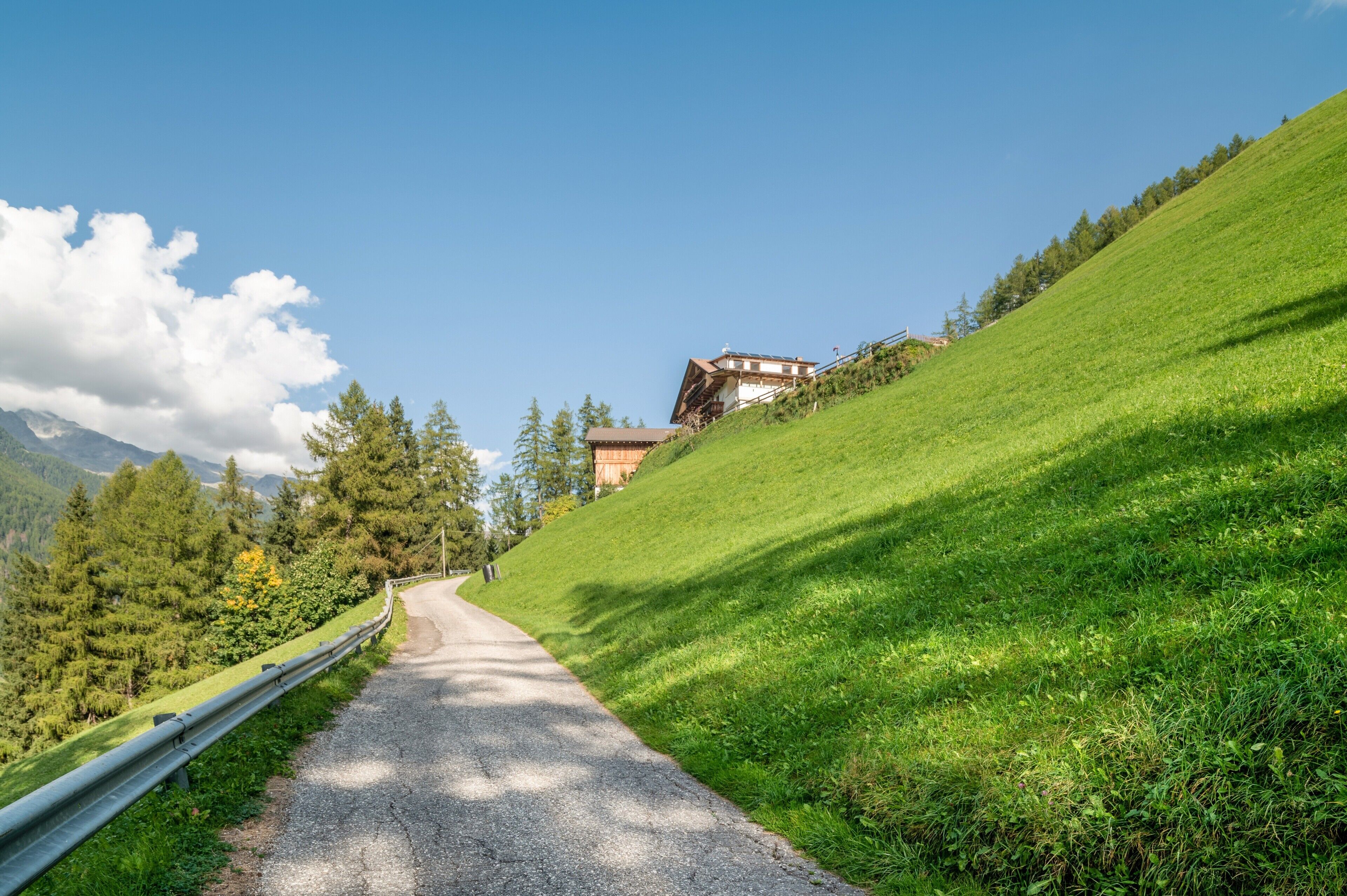 Une route asphaltée bordée d’une barrière métallique, une colline avec un bâtiment, et une montagne boisée en arrière-plan.