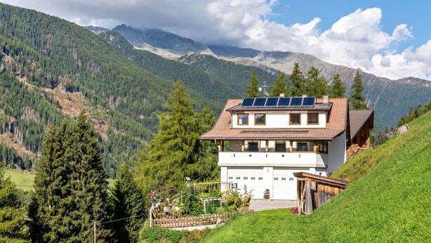 Una casa de dos pisos con paneles solares, rodeada de montañas y vegetación.