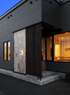 Un bâtiment moderne à la façade en briques sombres, doté d’une grande porte en bois avec le logo « M », et dont l’intérieur est visible, notamment un espace dédié à la salle à manger.