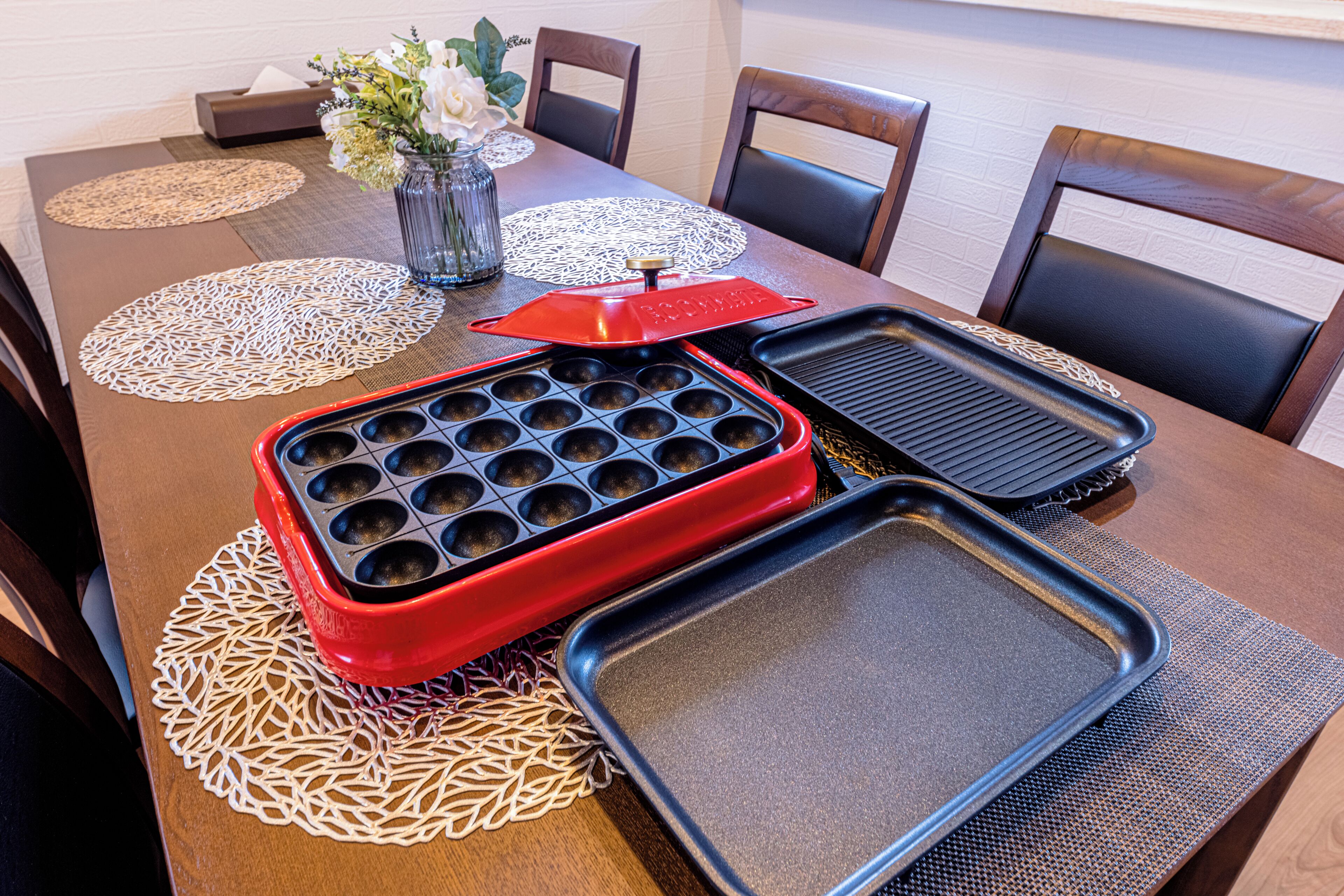 A dining table with a red waffle iron, two black baking trays, and a vase with white flowers.