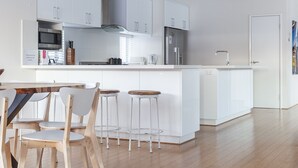 A modern kitchen with white cabinetry, a central island with stools, and a dining area with wooden chairs. - southbeach-house - Stylish & Spacious beach mansion (South Fremantle)