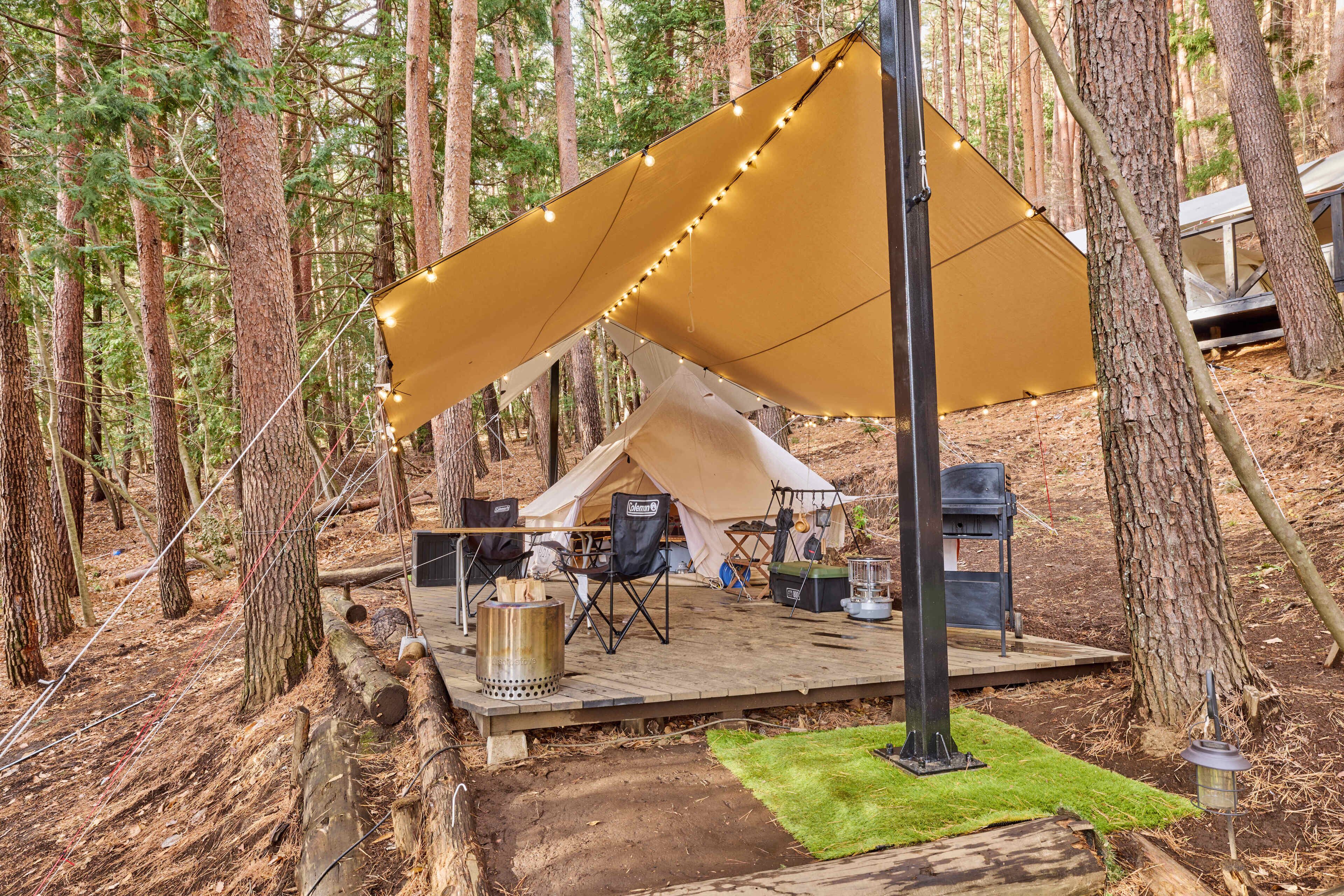 A forest campsite with a tent, deck, chairs, and a grill.