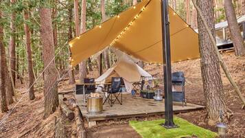 A forest campsite with a tent, deck, chairs, and a grill.