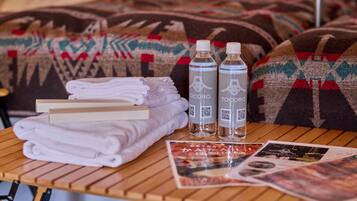 Un lit bien fait, recouvert d’une couverture à motifs, deux bouteilles d’eau Tocoro, des serviettes pliées et un magazine posés sur une table en bois.
