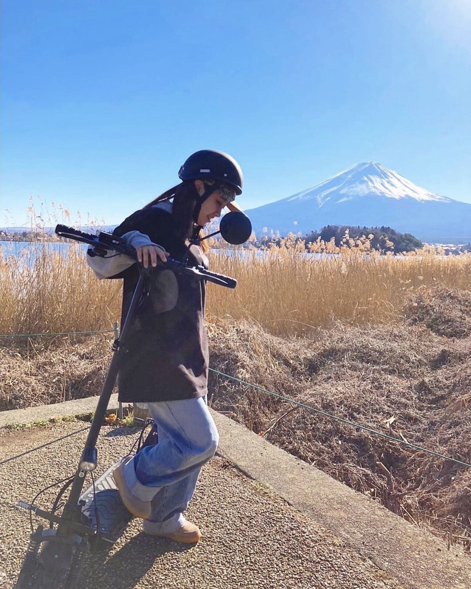 Eine Person fährt auf einem Scooter, trägt Helm und Mikrofon und steht auf einem Weg mit trockenem Gras, im Hintergrund ist eine Berglandschaft zu sehen.