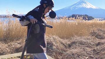 Eine Person fährt auf einem Scooter, trägt Helm und Mikrofon und steht auf einem Weg mit trockenem Gras, im Hintergrund ist eine Berglandschaft zu sehen.
