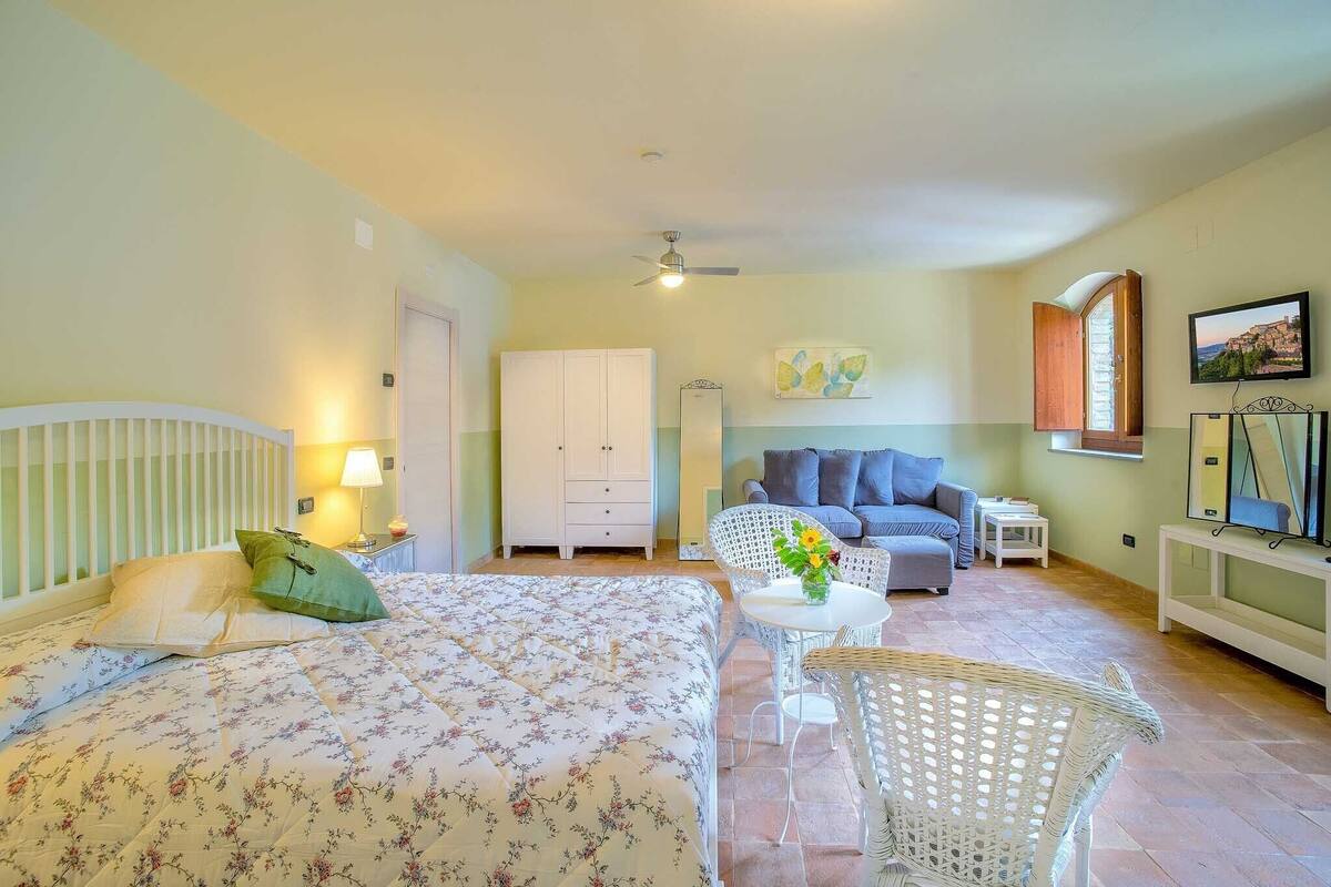 A bedroom with a bed, a wardrobe, a sofa, a small table with a vase, and a chair.