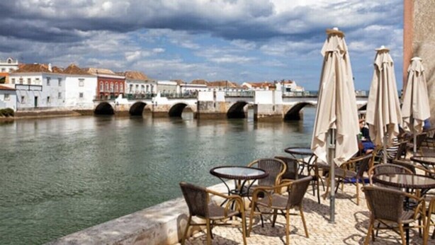 Un café situé au bord de la rivière, avec une terrasse, un pont en arrière-plan et des bâtiments qui bordent la rivière.