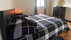 Ein Schlafzimmer mit Bett, Nachttisch, Lampe, Kommode und einem Fenster mit Vorhängen.