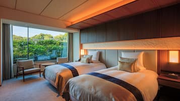 Habitación de hotel con dos camas, una zona de estar con una silla, una mesita y vistas a zonas verdes.