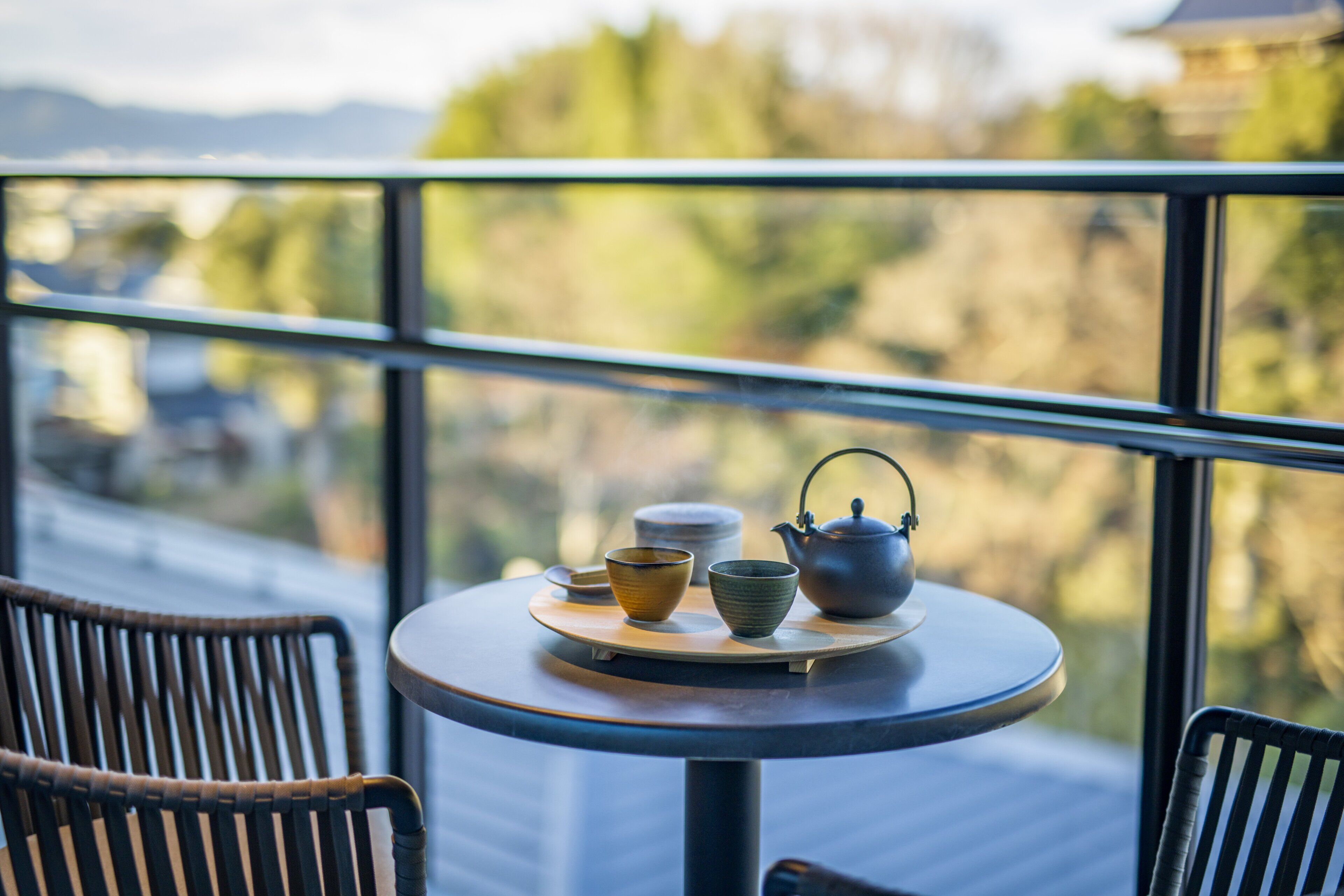 A round table with a teapot, cups, and bowls on a balcony overlooking a scenic view.