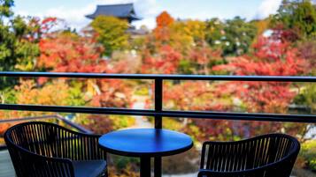 A balcony with two black chairs and a small round table, overlooking a garden with autumn-colored trees and a traditional building in the background.