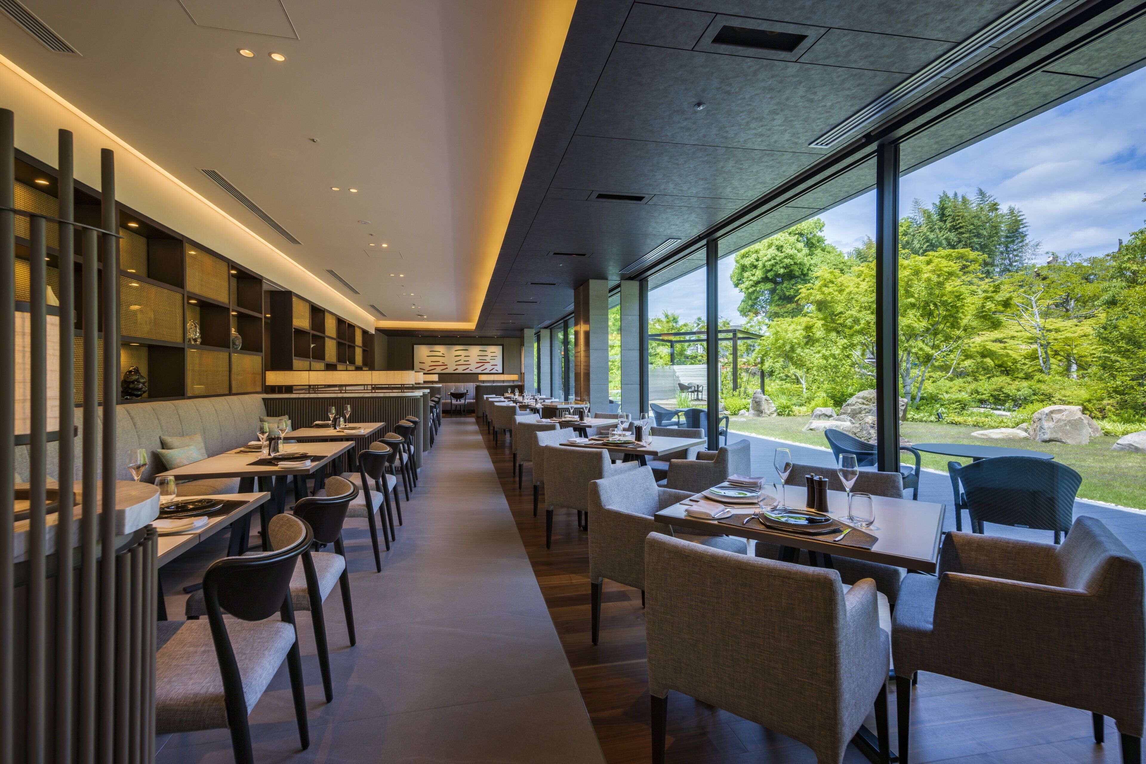 A modern restaurant with long tables and chairs, a bar area, and large windows offering a view of the outdoors.