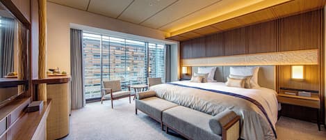 Habitación de hotel moderna con una cama grande, zona de estar, escritorio y vistas a la ciudad a través de amplios ventanales.