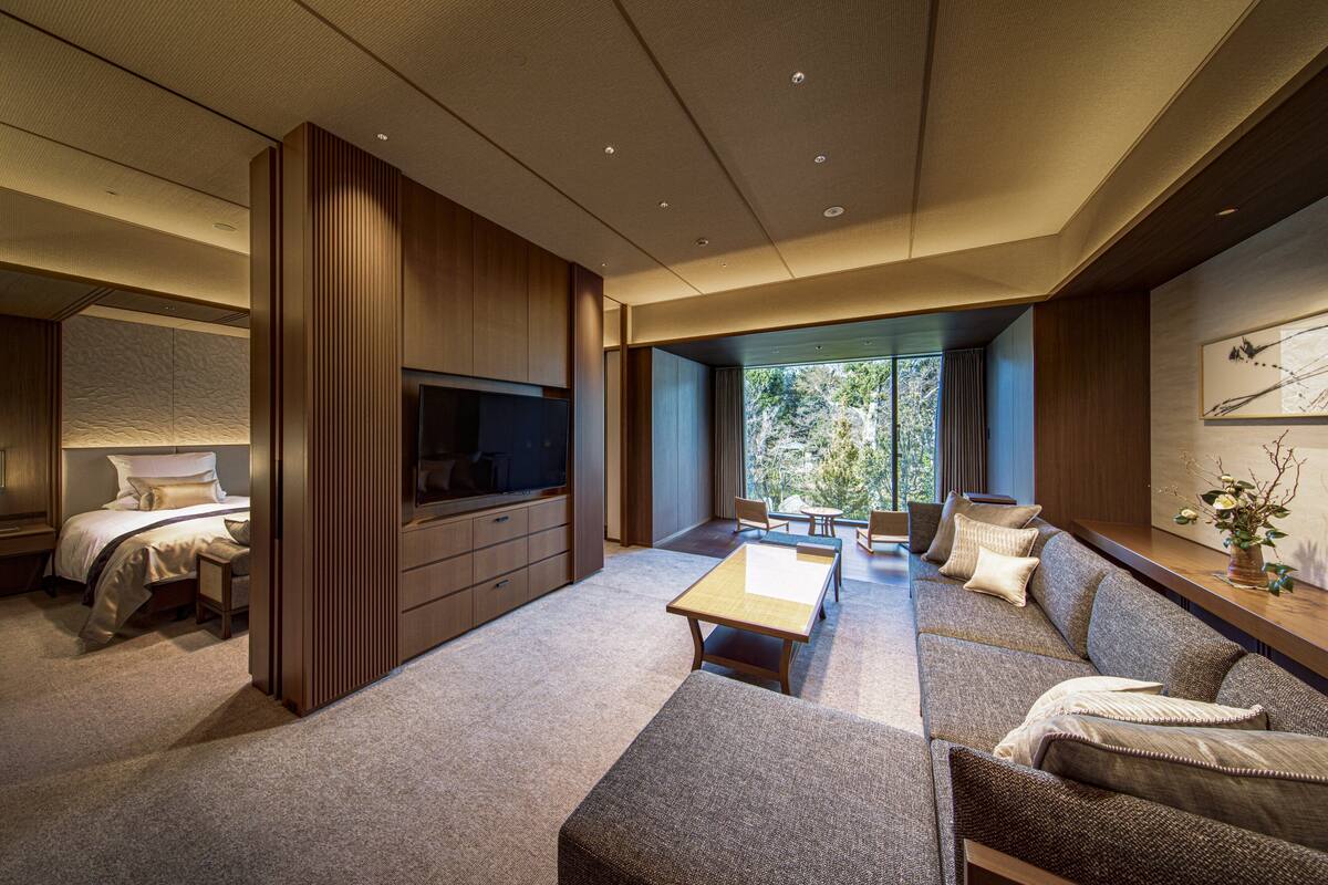 A modern hotel room with a large sofa, a flat-screen TV, a dining table, and a large window offering a view of the outdoors.
