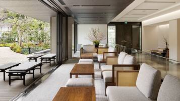 A modern indoor seating area with wooden tables and cushioned chairs, large windows offering a view of a landscaped garden.