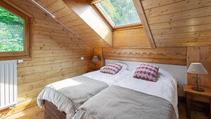 A cozy wooden cabin bedroom with a skylight, a bed with two pillows, a bedside table with a lamp, and a radiator.