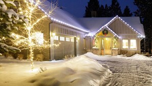 Un edificio de dos pisos con techo nevado, un garaje y un porche delantero adornado con luces y un adorno navideño.
