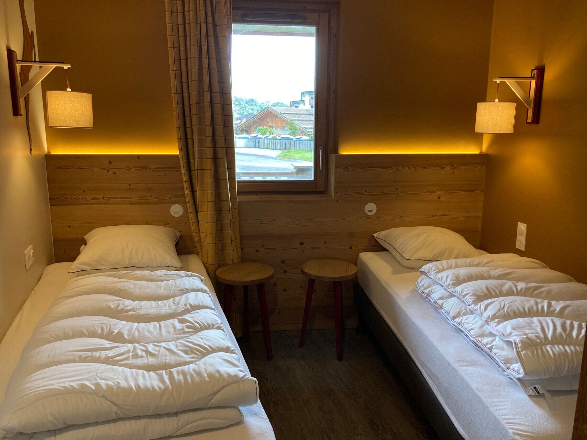 Une chambre avec deux lits simples, une petite fenêtre et une vue sur un bâtiment à l’extérieur.