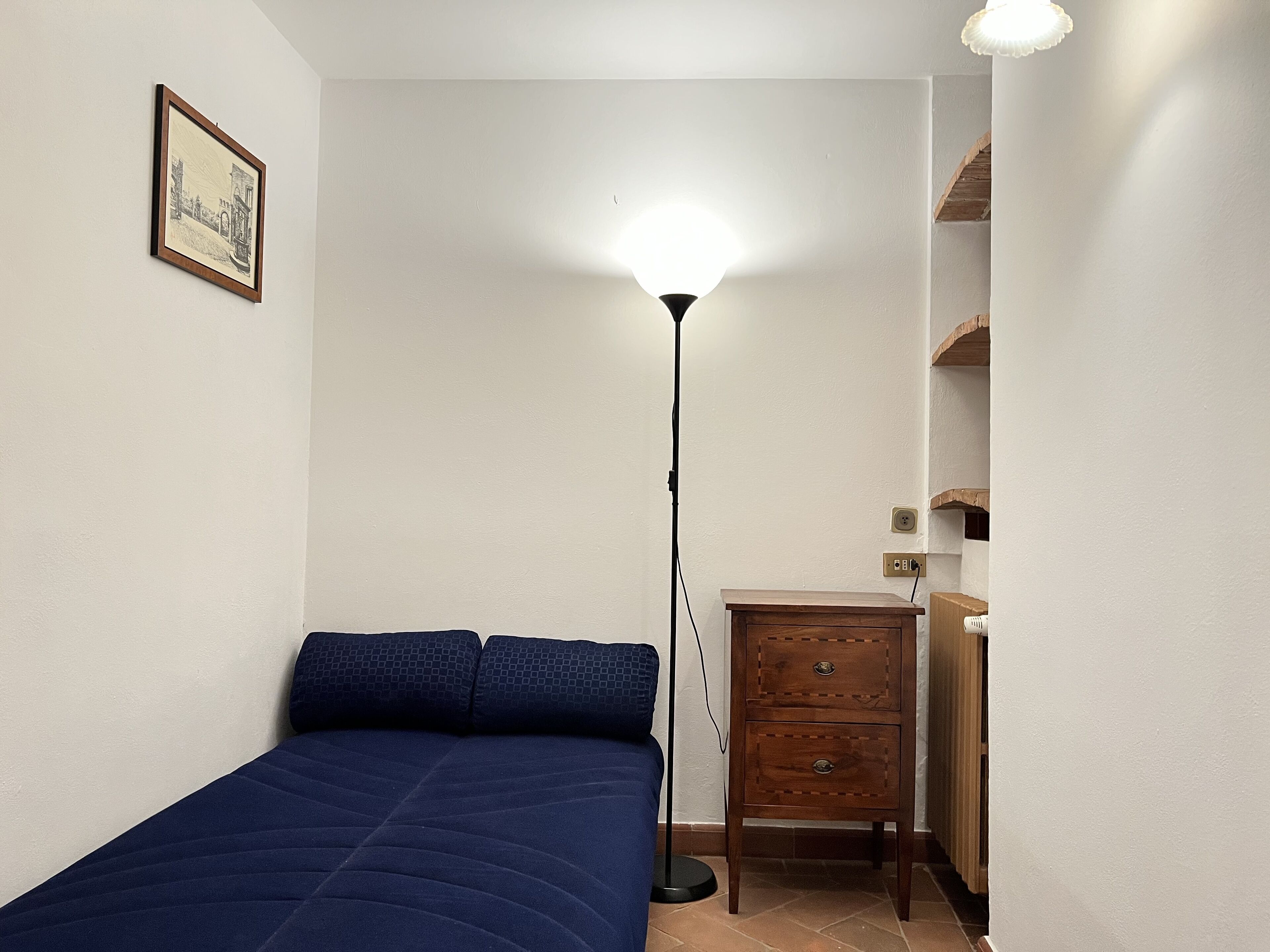 A bedroom with a bed, a bedside table, a floor lamp, and a small wooden cabinet.