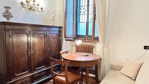 A room with a wooden cabinet, a round wooden table with a chair, a radiator, a sofa, and a window with curtains. - Duprè Apartment (Siena)