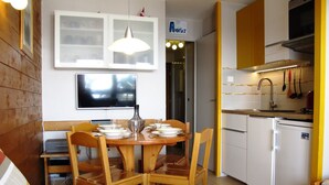 A dining area with a wooden table and chairs, a television mounted above the table, and a kitchen with white cabinets and a stove. - Renovated studio with south-facing balcony in Avoriaz (Avoriaz)