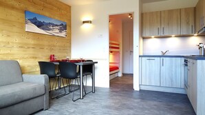 A modern room with a wooden wall, a dining table with chairs, a sofa, a kitchen area with cabinets, and a bed with red bedding. - Renovated studio in Avoriaz center for 4 pers. (Avoriaz)