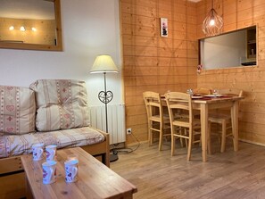 Um quarto aconchegante com piso de madeira, um sofá com almofadas estampadas, uma mesa de jantar com cadeiras, um abajur e um espelho na parede.