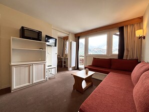 A hotel room with a red sofa, a wooden coffee table, a TV on a shelf, and a balcony with a view.