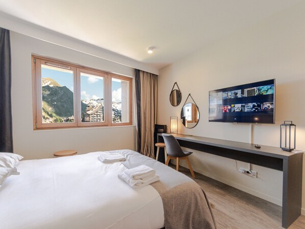A hotel room with a bed, a desk, a chair, a TV, and a window with a mountain view.