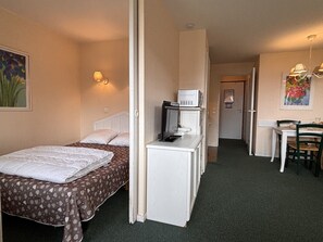 Ein kompaktes Hotelzimmer mit einem Bett, einem Schreibtisch mit Computer und Mikrowelle, einem Essbereich mit Tisch und Stühlen sowie einem Flur, der zu einem weiteren Zimmer führt.