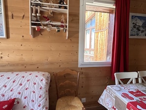 A wooden cabin room with a bed, a chair, a shelf with decorations, and a window with a view of a wooden structure.