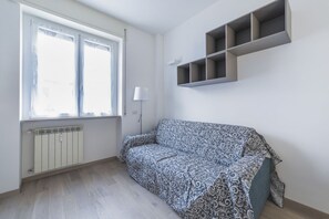 Una habitación con un sofá estampado, un radiador, una ventana con cortinas y un estante fijado a la pared.