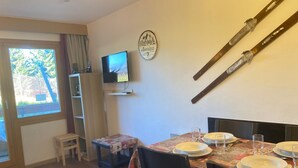 A dining area with a table set for four, a television on a shelf, and a window with curtains.