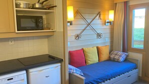A compact room with a kitchenette, a bed with colorful pillows, and a window with curtains.
