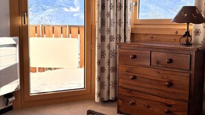 Une pièce avec une commode en bois, un bureau avec une lampe et une vue sur un paysage montagneux enneigé, visible à travers une grande fenêtre.