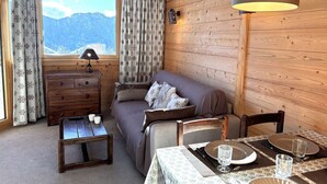 Une pièce chaleureuse avec un canapé, une commode en bois, une table dressée pour le repas et une fenêtre avec des rideaux.