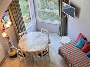 A cozy room with a round table, two chairs, a bed with colorful pillows, a mounted TV, and a window with curtains.